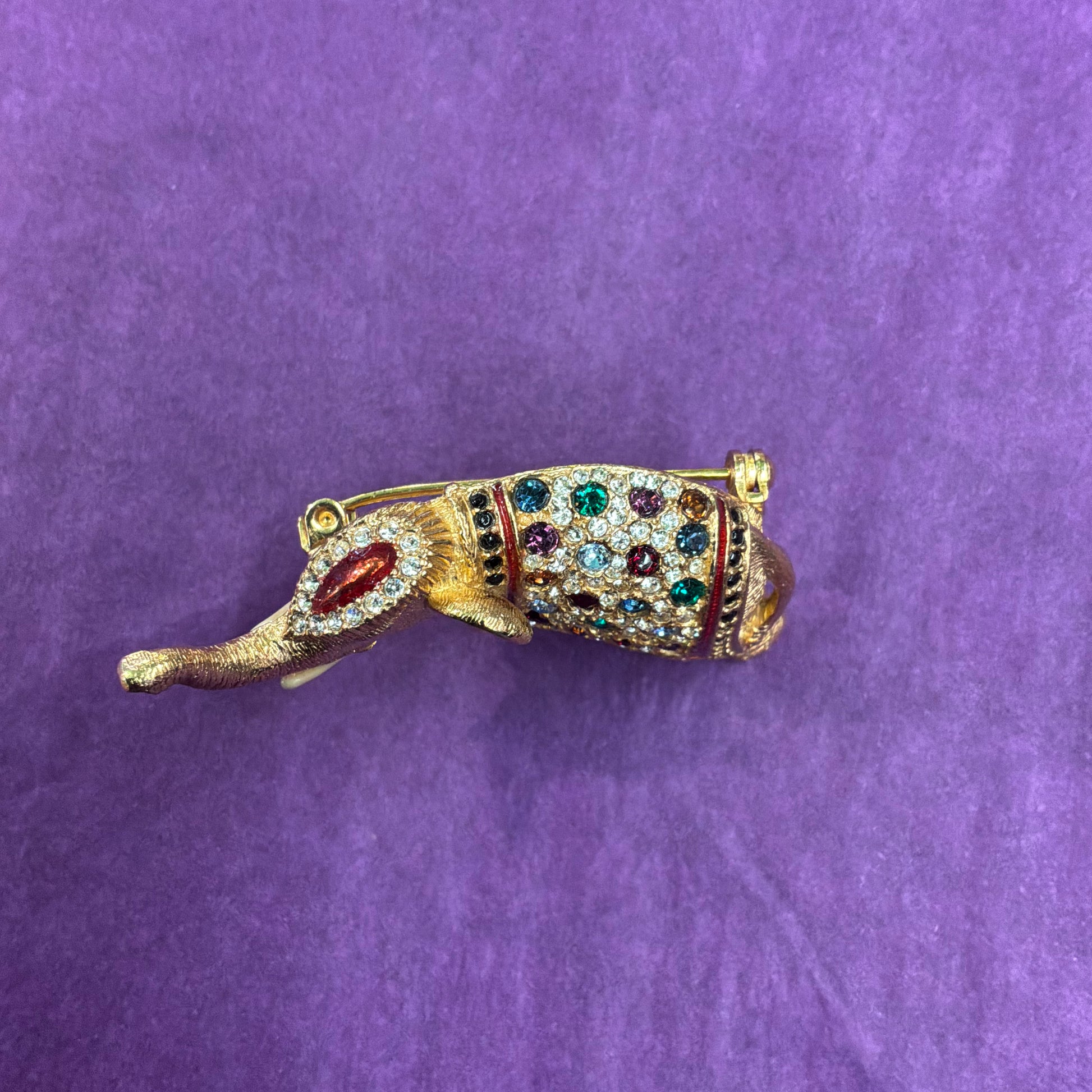 A gold-toned elephant-shaped brooch decorated with clear rhinestones and colorful gemstones in green, red, and blue, set against a textured purple background.